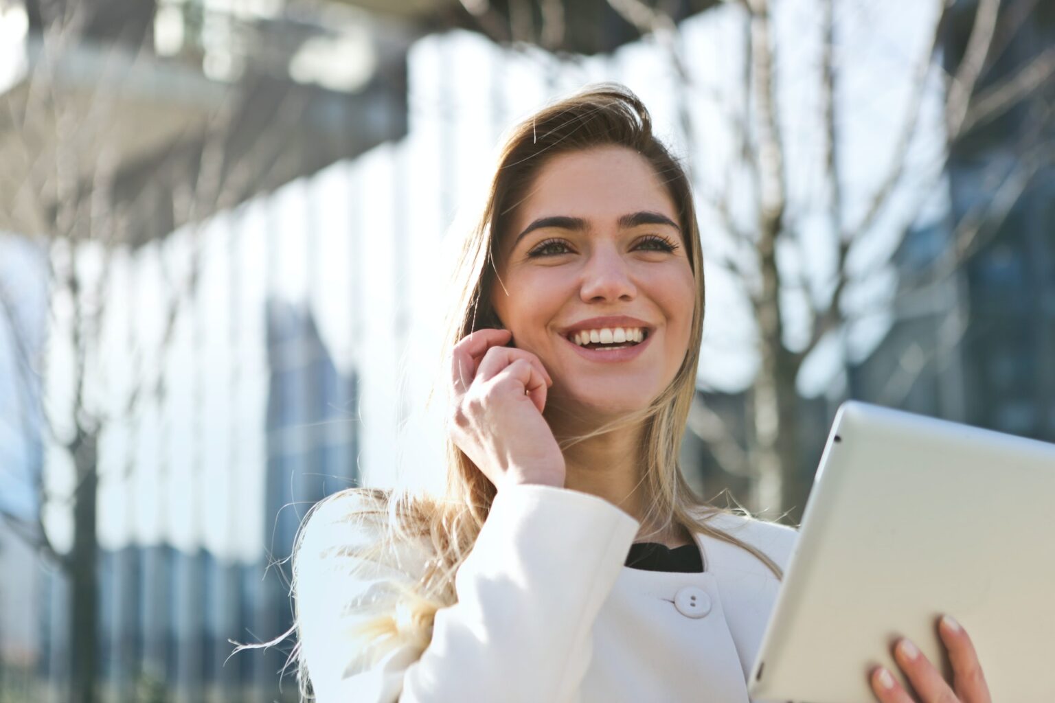 A Caucasian woman with blonde hair is holding her cell phone in one hand and a tablet in the other hand. She's wearing a white sweater, and she's standing outside in front of a fence and a leafless tree.